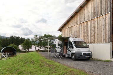 Ein Wohnmobil steht mit ausgefahrener Markise und Stühlen auf einem Stellplatz. Im Hintergrund stehen Alpakas. | © Bodenmais Tourismus & Marketing GmbH