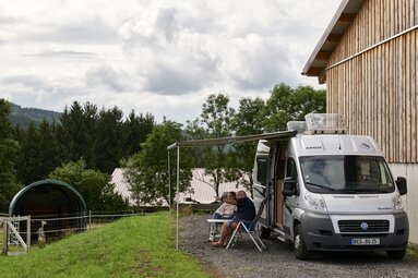 Vor einer Wiese mit Alpakas steht ein Wohnmobil. Unter der Markise sitzen zwei Personen, die sich unterhalten und in die Ferne zeigen. | © Bodenmais Tourismus & Marketing GmbH