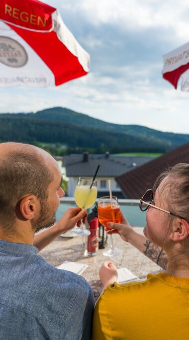 Blick von hinten auf ein Paar, das sich zuprostet. Vor ihnen liegt eine tolle Aussicht die von weiß-rot gestreiften Sonnenschirmen umrahmt wird. | © Bodenmais Tourismus & Marketing GmbH