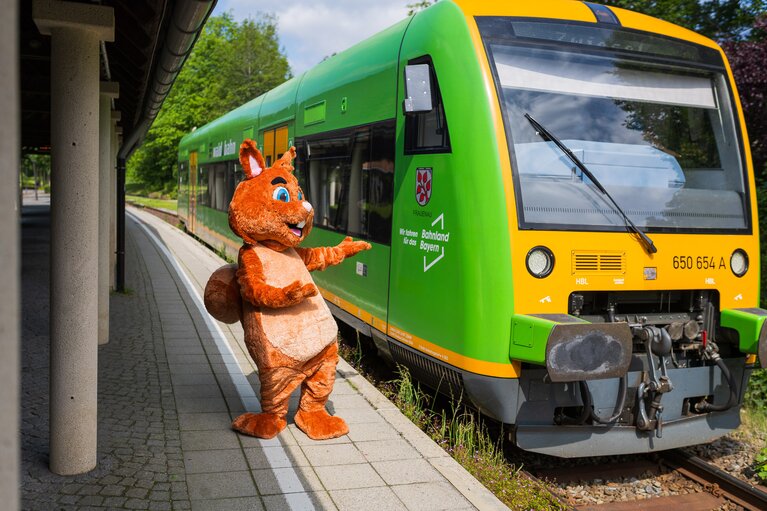 Das Bodenmais Maskottchen Eichhörnchen Edi zeigt auf der grüne Waldbahn, die neben ihm am Gleis angehalten hat. | © Bodenmais Tourismus & Marketing GmbH