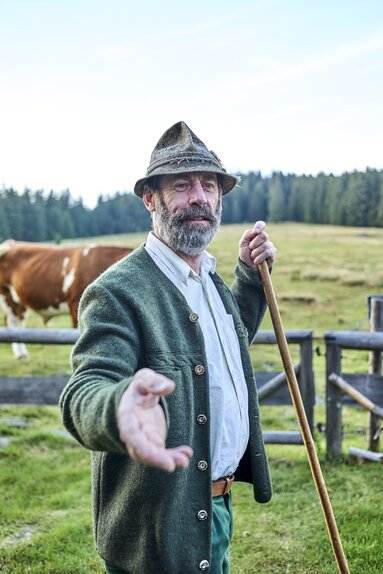 Porträts eines Mannes mit grauem Bart, Trachtenhut und -jacke sowie Wanderstock auf einer Kuhweide. | © Woidlife 