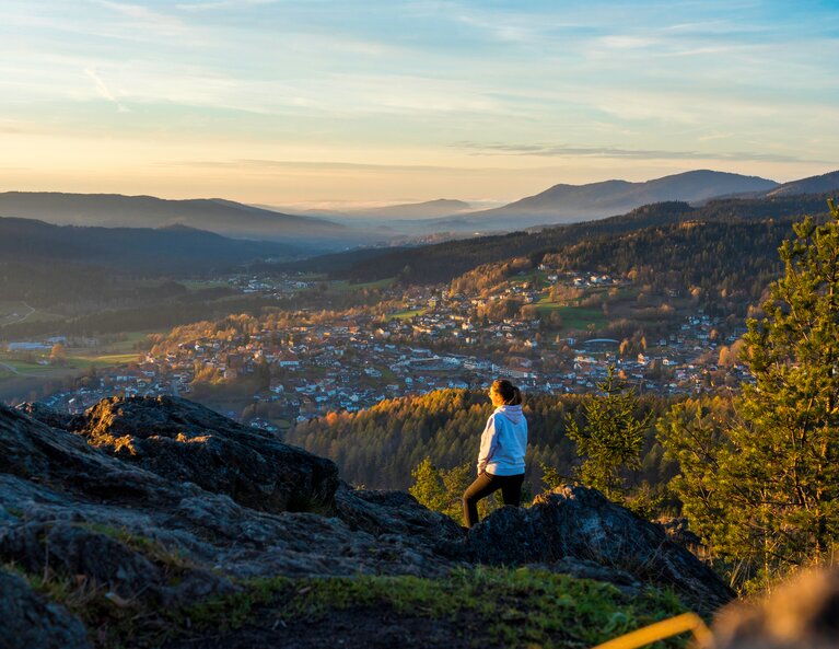 Ein Frau steht mit dem Rücken zur Kamera und blickt vom Silberberg aus hinab nach Bodenmais. Das herbstliche Tal wird von der untergehenden Sonne angestrahlt. | © Bodenmais Tourismus & Marketing GmbH